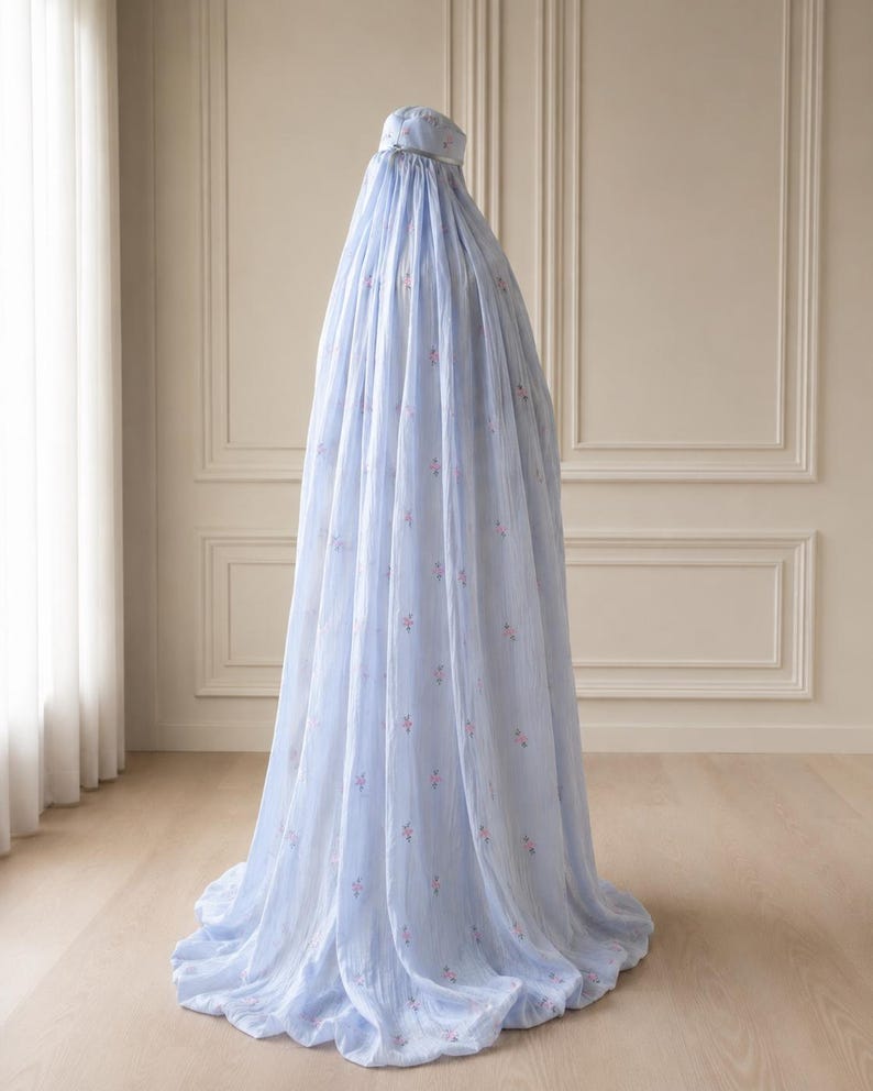 May include: A light blue, floor-length prayer garment featuring a matching head covering and a subtle, repeating pattern of small pink floral motifs. The fabric is lightweight and drapes fully to the floor, creating a voluminous silhouette.