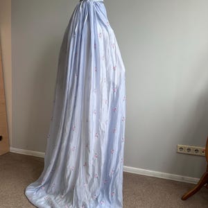 May include: A light blue, floor-length garment with a subtle vertical texture and a repeating pattern of small pink floral clusters. The piece features a gathered neckline with a matching light blue ribbon tie and a loose, flowing silhouette.