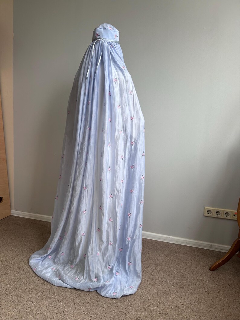 May include: A light blue, floor-length garment with a subtle vertical texture and a repeating pattern of small pink floral clusters. The piece features a gathered neckline with a matching light blue ribbon tie and a loose, flowing silhouette.