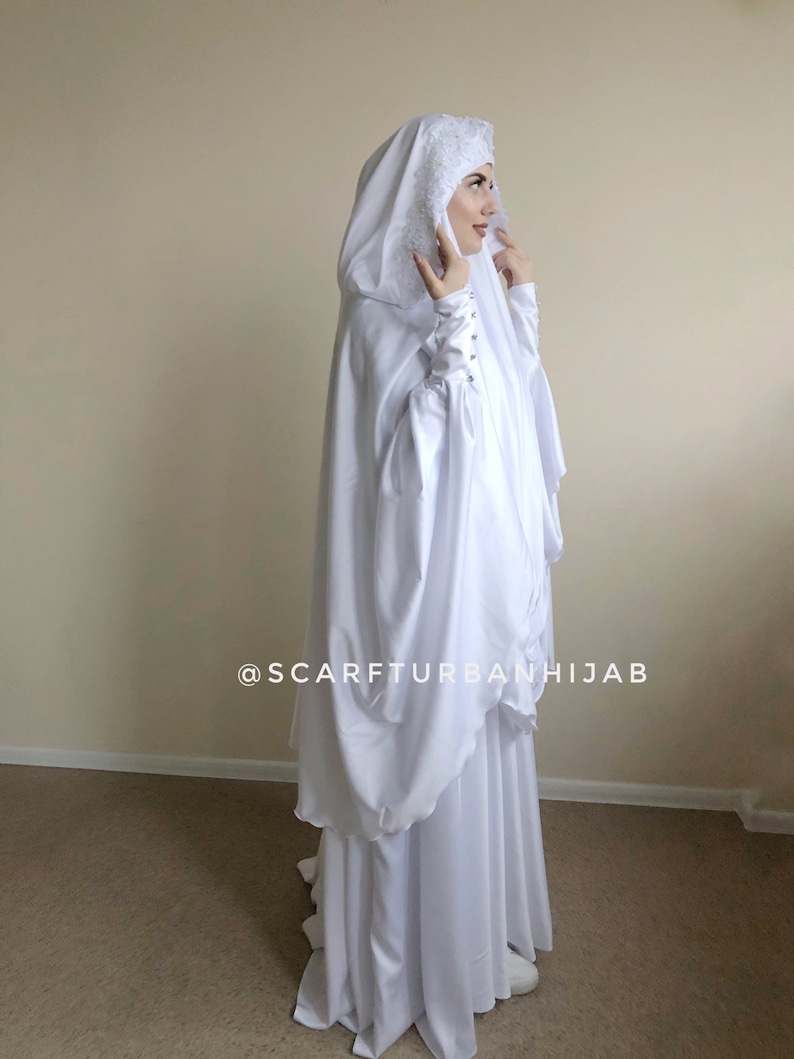 May include: White prayer shawl with a lace trim and a hood. The shawl has a gathered design and is worn by a person. The text "@scarfturbanhijab" is visible on the shawl.