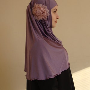 May include: A woman wearing a purple hijab with a pink flower accent. The hijab is made of a soft, flowing fabric. She is wearing a black abaya.
