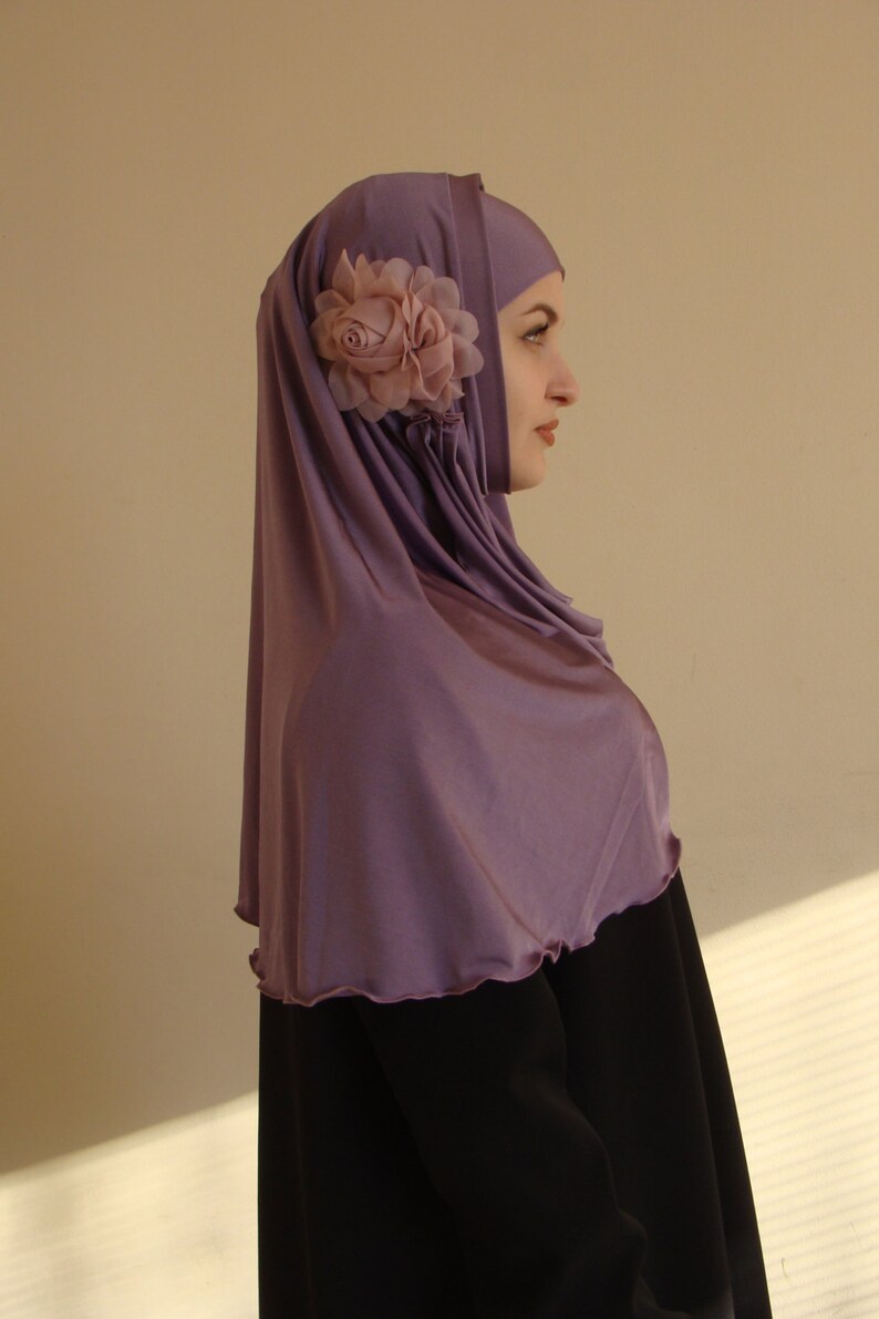 May include: A woman wearing a purple hijab with a pink flower accent. The hijab is made of a soft, flowing fabric. She is wearing a black abaya.
