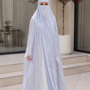 May include: A light blue, floor-length, sheer, layered abaya with a matching niqab and head covering. The fabric features a delicate, scattered floral pattern in soft pink and white tones, draping over a solid white, long-sleeved undergarment.