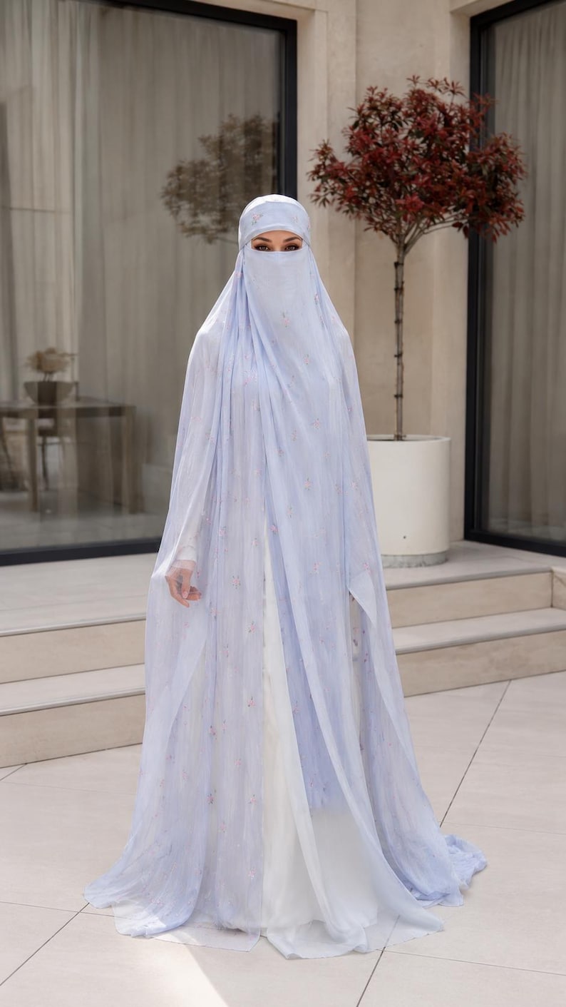 May include: A light blue, floor-length, sheer, layered abaya with a matching niqab and head covering. The fabric features a delicate, scattered floral pattern in soft pink and white tones, draping over a solid white, long-sleeved undergarment.