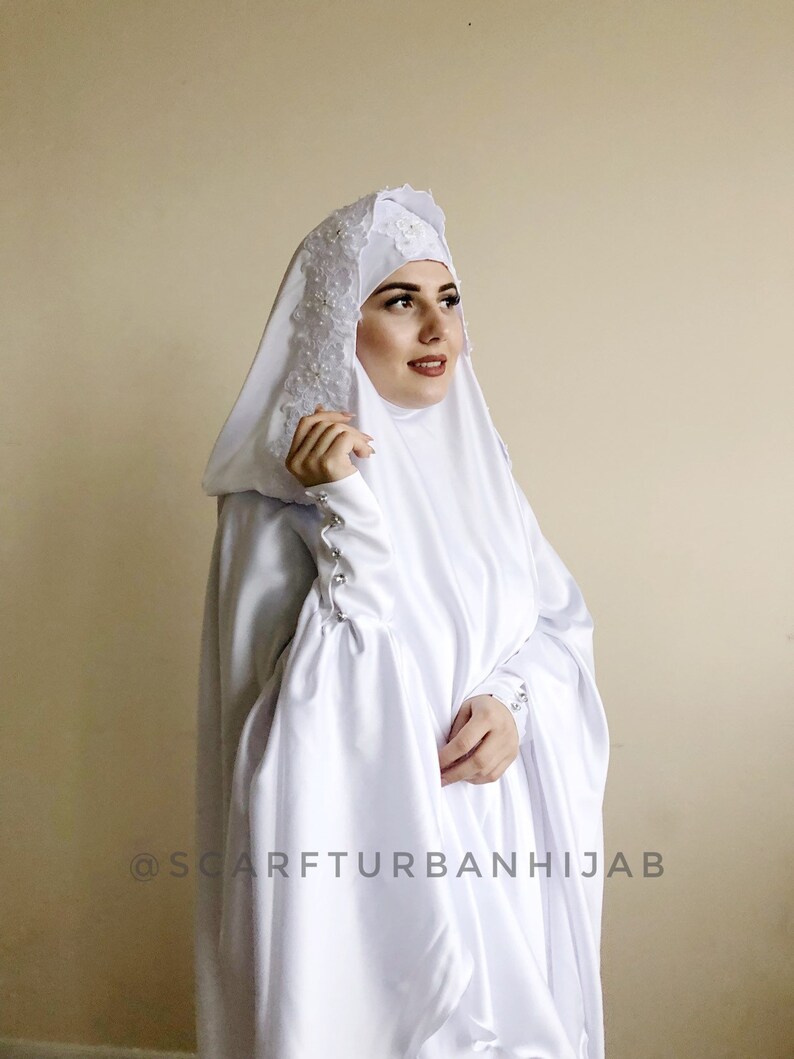 May include: White satin hijab with a lace trim and pearl embellishments. The hijab has a hood and long sleeves with button closures. The text "@scarfturbanhijab" is visible at the bottom of the image.