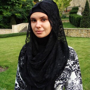 May include: A woman wearing a black lace hijab over a white and black patterned dress. The dress has a floral design.