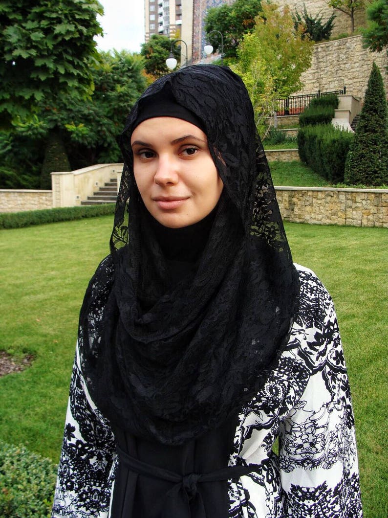 May include: A woman wearing a black lace hijab over a white and black patterned dress. The dress has a floral design.
