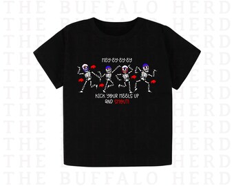 May include: A black short-sleeved t-shirt featuring a graphic of four dancing skeletons wearing blue and red hats. The text reads HEY-EY-EY-EY and KICK YOUR HEELS UP AND SHOUT! with small red buffalo silhouettes interspersed among the skeletons.