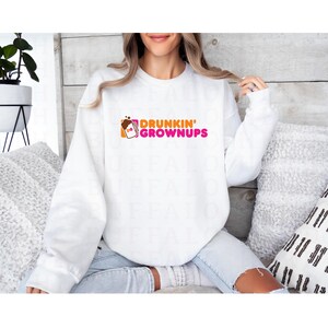 K&ouml;nnte beinhalten: Wei&szlig;er Pullover mit einer pinkfarbenen und orangenen Grafik, auf der "Drunkin' Grownups" steht, mit einem stilisierten Bild einer Kaffeetasse.