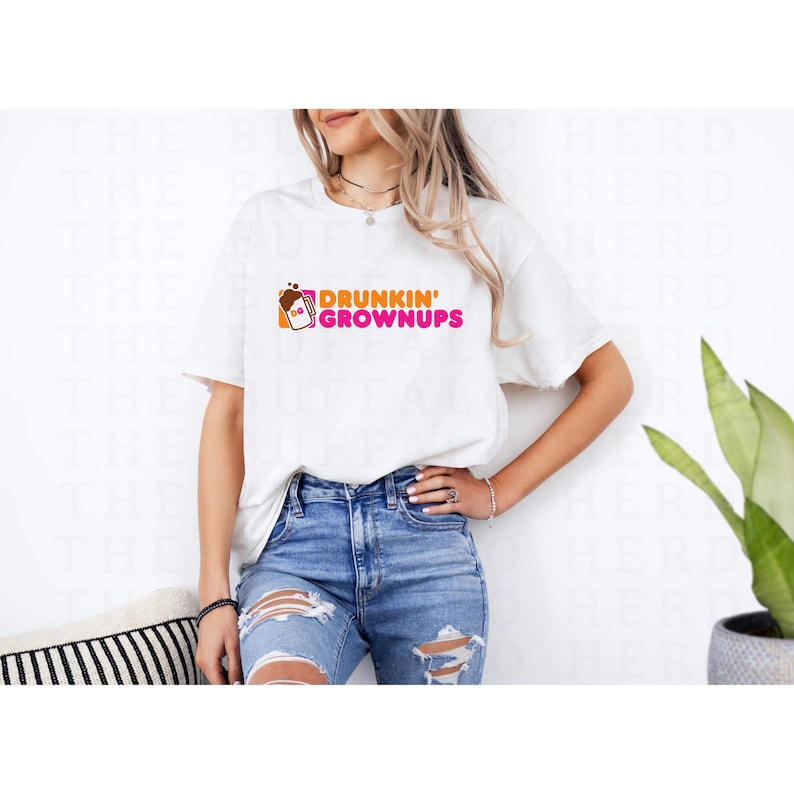 May include: A white t-shirt with the words 'DRUNKIN' GROWNUPS' printed in pink and orange. The shirt is being worn by a person with blonde hair and blue jeans.