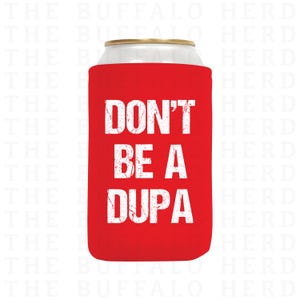 May include: A red can cooler with the text "DON'T BE A DUPA" in white, distressed lettering. The cooler is designed to fit around a standard beverage can, keeping drinks cold. The background is white.