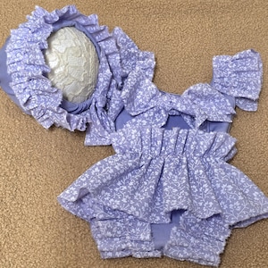 May include: A baby outfit set in a light purple floral print. The set includes a bonnet with a ruffled brim, a top with ruffled sleeves and a skirt with multiple layers of ruffles. The outfit is made of a soft fabric and is designed for infants.