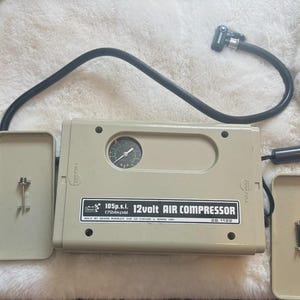 Könnte beinhalten: Beiger 12-Volt-Luftkompressor mit schwarzem Schlauch und Manometer. Der Kompressor trägt die Aufschrift "105 p.s.i. 12 volt AIR COMPRESSOR". Zwei beige Abdeckungen sind geöffnet und geben die Innenteile frei. Für den Einsatz im Automobilbereich konzipiert.