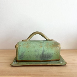 May include: A handmade ceramic butter dish with a rectangular base and a domed lid. The dish is glazed in shades of green and brown, with a curved handle on top. The butter dish sits on a matching rectangular plate.