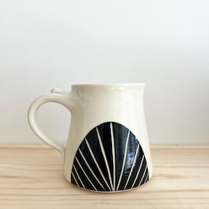 May include: A white ceramic mug with a black geometric design. The design features a half-circle with radiating lines.