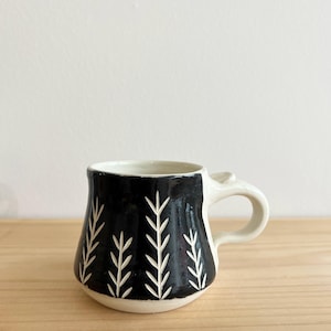 May include: A handmade ceramic mug with a black exterior and white leaf-like designs. The mug has a white handle and rim, and is set against a neutral background. This unique mug is perfect for coffee or tea.