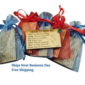 May include: A close-up of a mood ring in a small, red, organza bag. The ring is surrounded by other bags in various colors. A card with the text "ENCHANTED MOOD RING" and a color chart is visible. The card lists the different colors of the ring and their corresponding moods.