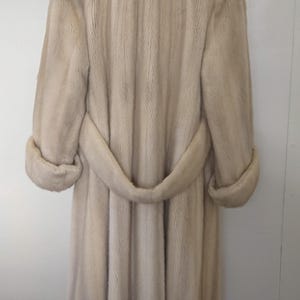 May include: A long, light beige fur coat with a belt. The coat has a classic design and is made of soft, luxurious fur.