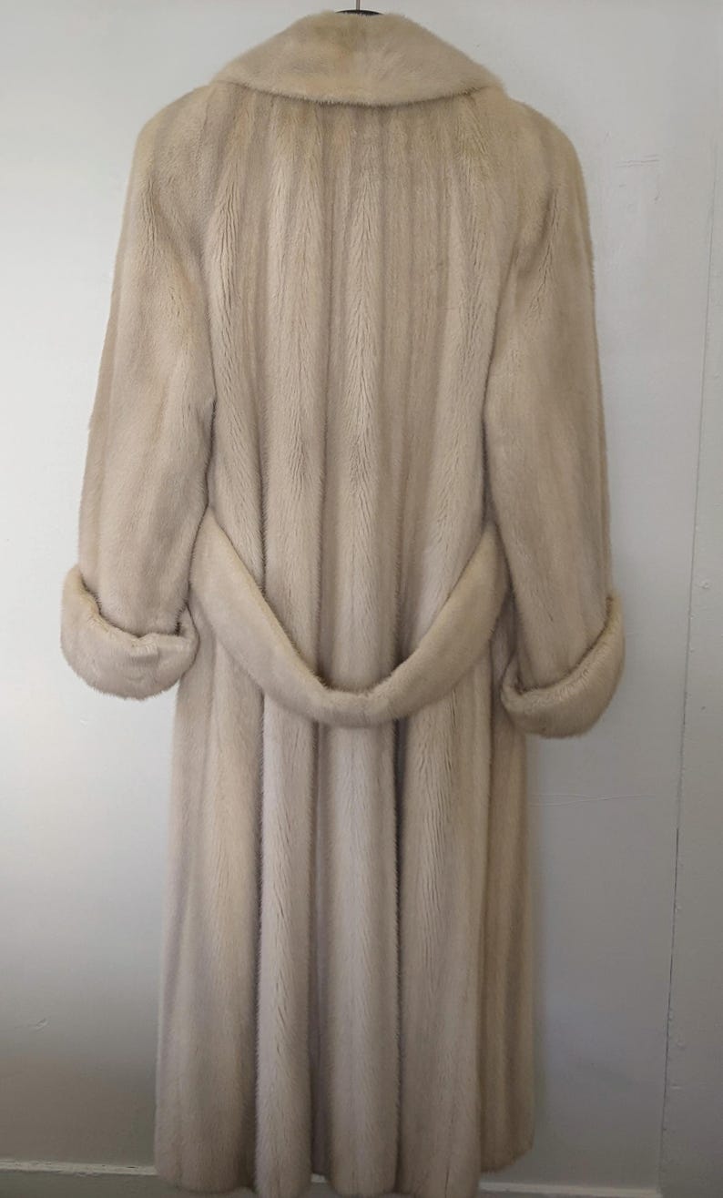 May include: A long, light beige fur coat with a belt. The coat has a classic design and is made of soft, luxurious fur.