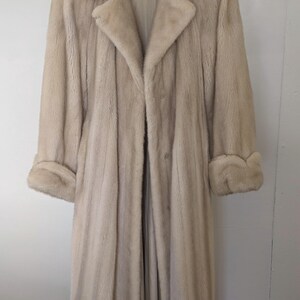 May include: A long, light beige fur coat with a collar and cuffs. The coat is hanging on a hanger.