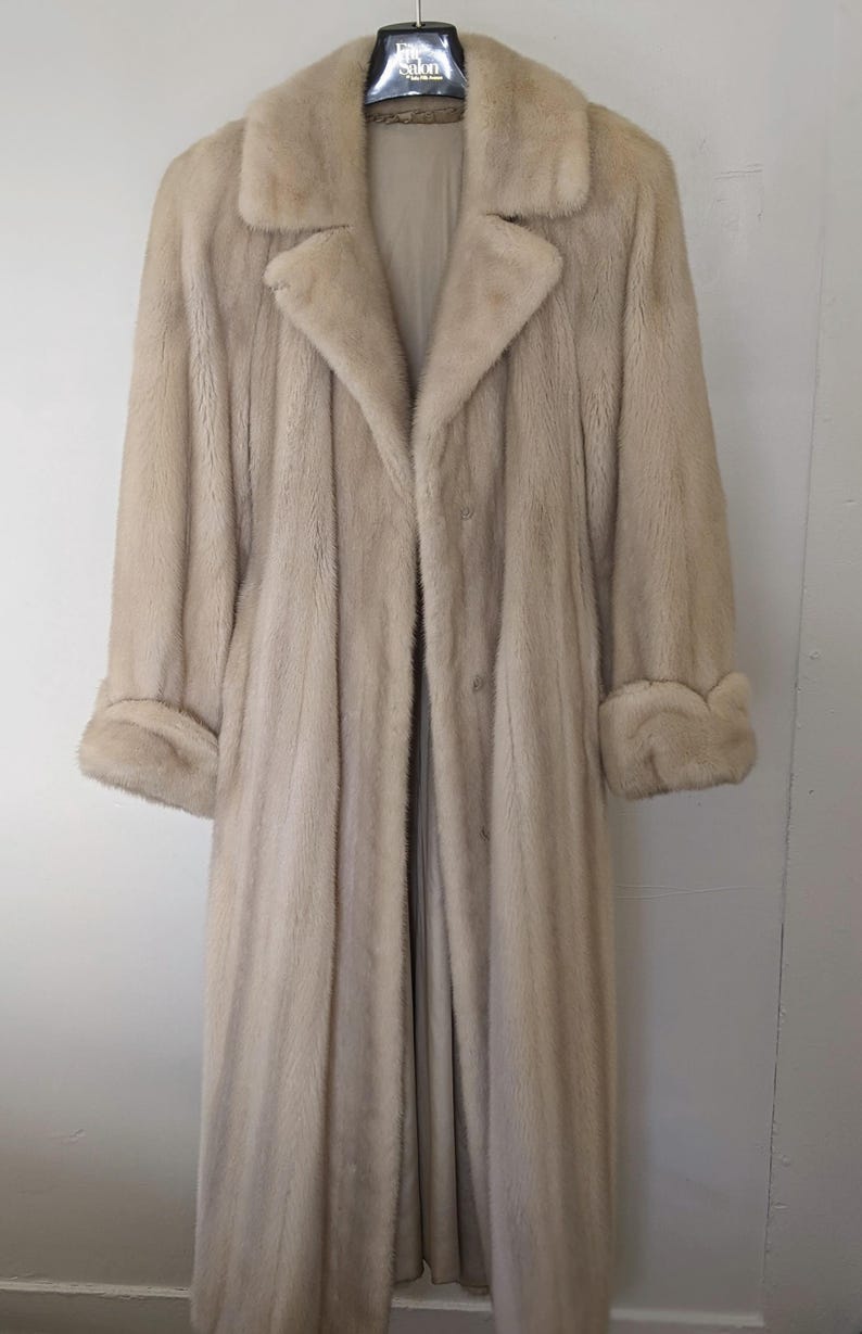 May include: A long, light beige fur coat with a collar and cuffs. The coat is hanging on a hanger.