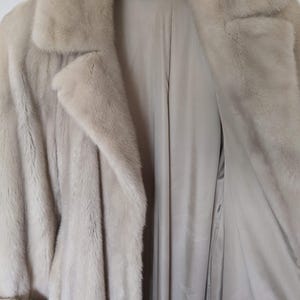 May include: A light beige fur coat with a long, flowing design. The coat has a collar and a button closure. The lining of the coat is a light beige color.