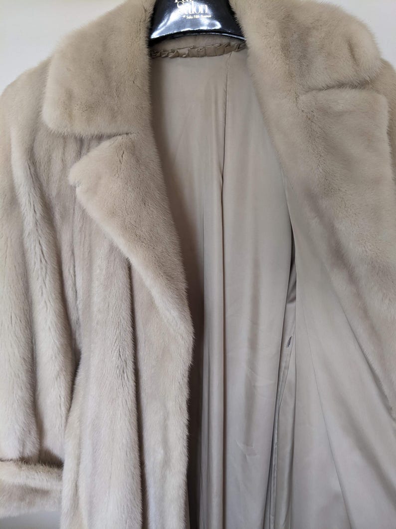 May include: A light beige fur coat with a long, flowing design. The coat has a collar and a button closure. The lining of the coat is a light beige color.