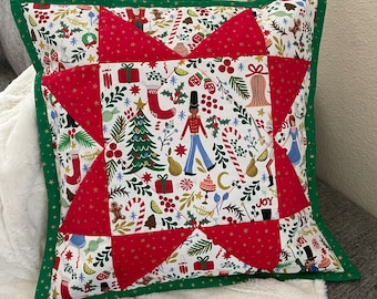 May include: A red and green Christmas pillow with a star design. The pillow is made of fabric with a white background and a variety of Christmas-themed patterns, including a Christmas tree, a nutcracker, and candy canes. The pillow has a green border.