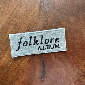 May include: A white embroidered patch with the text "folklore ALBUM" in black lettering.