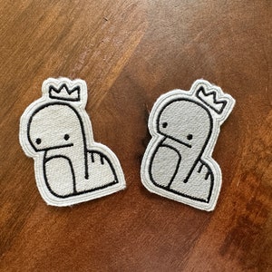 May include: Two white embroidered iron-on patches of a cartoon character wearing a crown. The character is a simple line drawing of a creature with a crown on its head.