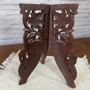 May include: A dark brown, intricately carved wooden stand with a three-legged base. The stand features leaf and floral cutouts on the sides and is displayed on a white, textured surface. The stand is approximately 10 inches tall.