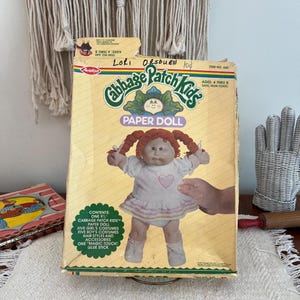 May include: A vintage Cabbage Patch Kids Paper Doll box. The box features a doll with red pigtails, wearing a white dress with a pink heart. The box reads "Cabbage Patch Kids Paper Doll" and includes the text "Ages 4 thru & Safe, Non-Toxic."