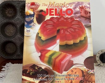 Op de afbeelding: Een kookboek getiteld "The Magic of JELLO" met afbeeldingen van kleurrijke gelatinedesserts. De omslag toont een gelaagde gelatinevorm en andere Jell-O-recepten. Het boek viert 100 jaar Jell-O.