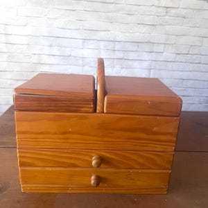 May include: A wooden sewing box with a hinged lid and a handle. The box has two drawers with round wooden knobs. The wood has a natural, light brown finish. The box is sitting on a wooden surface.
