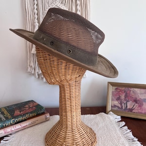 May include: A brown mesh-top hat with a wide brim, displayed on a wicker head form. The hat features a dark green band with two eyelets. The hat's brim and band are a darker shade of brown. The hat is a stylish accessory.