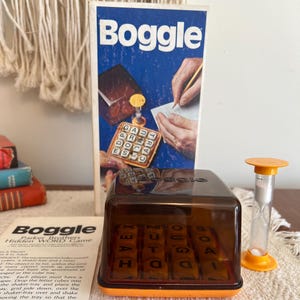Op de afbeelding: Een vintage Boggle bordspel met een bruine plastic dobbelsteenbak, een doorzichtig plastic deksel en een oranje basis. De dobbelsteenbak bevat 16 letterdobbelstenen. Het spel bevat ook een bruine doos met een blauwe achtergrond en het woord "Boggle" in witte letters. Er is ook een klein zandloper met een gele bovenkant en een witte basis.