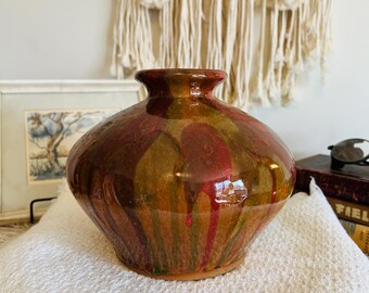 May include: A handcrafted ceramic vase with a glossy finish. The vase features a bulbous shape with a narrow neck and is adorned with a blend of red, green, and gold hues. The colors drip down the sides, creating a unique, artistic design.