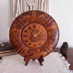 May include: A round, wooden clock with intricate carvings of animals and Mayan symbols. The clock features a cheetah, deer, monkey, and owl. The clock is displayed on a wooden stand.