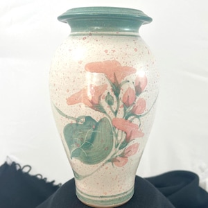 May include: A ceramic vase with a speckled cream background and a green rim. The vase features a floral design with pink flowers, green leaves, and buds. The vase is a decorative item for the home.