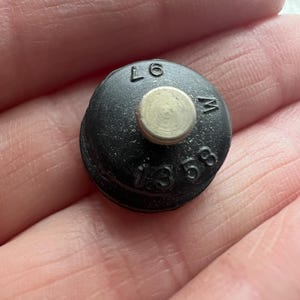 Könnte beinhalten: Schwarzer Plastikknopf mit einem silbernen Metallzentrum. Der Knopf hat die Buchstaben "L6" und "M" sowie die Nummer "1358" aufgedruckt.