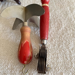 Peut inclure: Deux outils de cuisine vintage avec des manches en bois rouge. L'outil de gauche a trois lames et un crochet à l'extrémité du manche. L'outil de droite a une série de lames rondes.