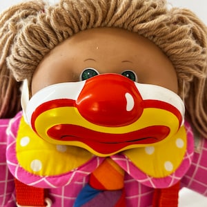 May include: A Cabbage Patch Kid doll wearing a clown costume with a red nose and a yellow and white mask. The doll has brown hair and green eyes. The doll is wearing a pink and white checkered shirt with a rainbow tie and white overalls.