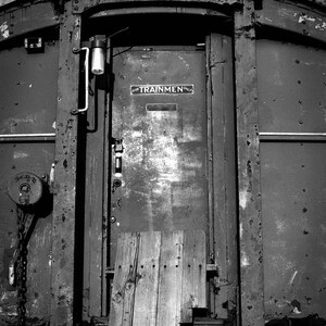 May include: A black and white photo of a weathered metal door with a sign that reads "For Trainmen Only". The door is on a train car and has a wooden plank on the bottom.