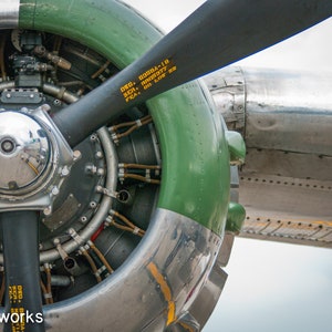 May include: Close-up of a vintage aircraft engine with a green and silver propeller. The engine has a silver casing with a green outer ring and a silver propeller blade. The engine has a series of pipes and wires connected to it. The propeller blade has the text "DRG - 2030A-10, S/N - 001777, P/EA - 001176-32C" written on it.