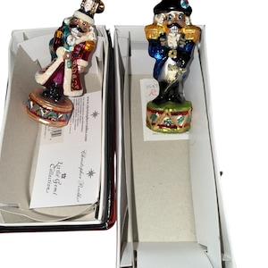 May include: Two glass Christmas ornaments shaped like nutcrackers, each in a box. One nutcracker wears a red coat and a white hat. The other wears a blue coat and a black hat. Both stand on a decorative drum base.