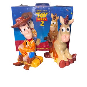 May include: A set of Toy Story 2 figurines featuring Woody and Bullseye, with a blue box in the background. Woody is in a cowboy hat and vest. Bullseye is a tan horse with red hooves. The box has the Disney Pixar Toy Story 2 logo.