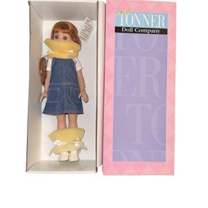 May include: A Tonner Doll Company doll in its original box. The doll has red hair, blue eyes, and is wearing a white shirt, denim jumper, and white shoes. The box is pink and features the Tonner Doll Company logo.