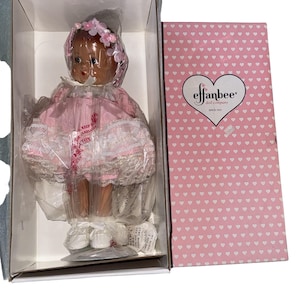 May include: A vintage Effanbee doll in its original box. The doll is dressed in a pink dress with white lace trim and a floral headpiece. The box is pink with a heart pattern and the Effanbee logo.