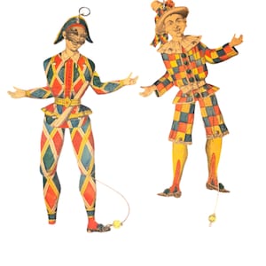 May include: Two vintage paper cutouts of Harlequin figures. The figures are dressed in colorful diamond-patterned costumes with hats and masks. Each figure has a string attached to a yellow ball at the bottom, and a metal ring at the top.