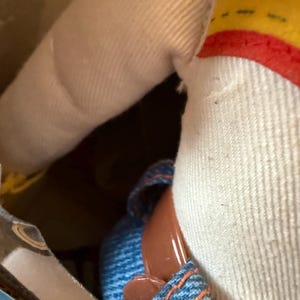 Puede incluir: Primer plano de una mu&ntilde;eca con una corona amarilla y roja, una camisa blanca y zapatos vaqueros azules con suelas marrones. Se ve el brazo de la mu&ntilde;eca, hecho de tela beige. La mu&ntilde;eca est&aacute; dentro de una caja de cart&oacute;n.